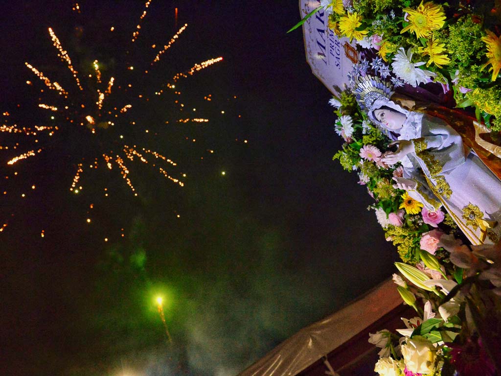 Toma en ángulo contrapicado que muestra en la esquina superior derecha la imagen iluminada de la Virgen rodeada de abundante arreglo floral con girasoles y rosas, y a la izquierda un espectáculo de pirotecnia dorada estallando contra el cielo nocturno.