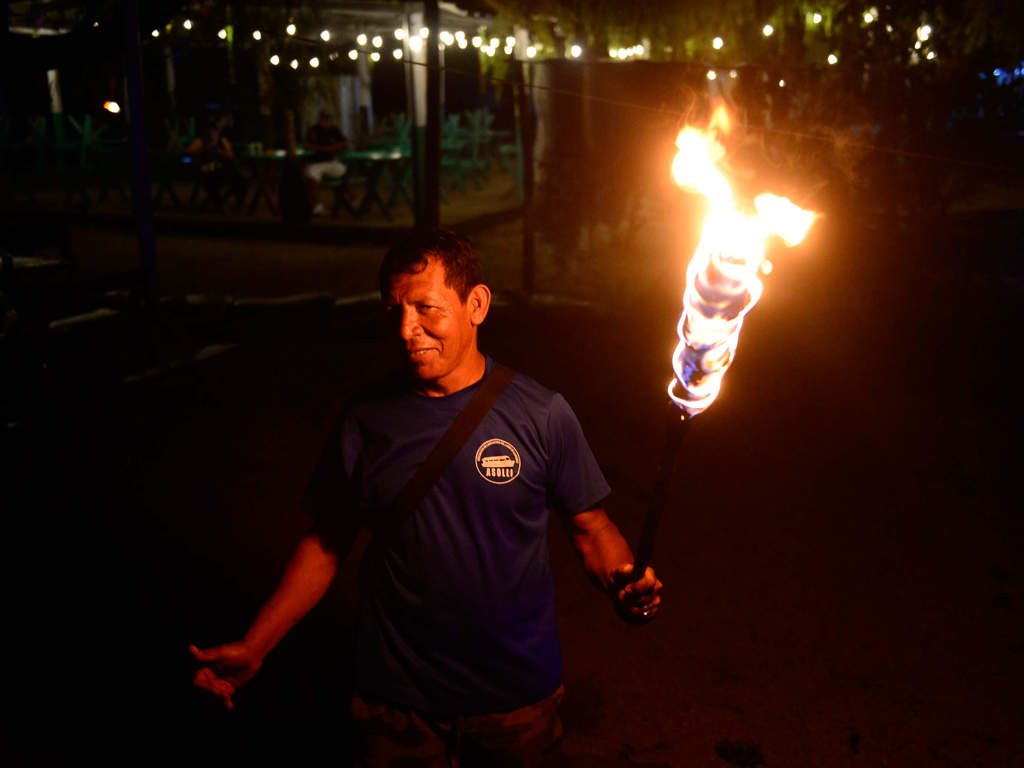 Un hombre sonriente con una camiseta azul que dice "ASOLLI" sostiene una gran antorcha encendida que ilumina su rostro y el área circundante por la noche. Al fondo se ven luces colgantes.