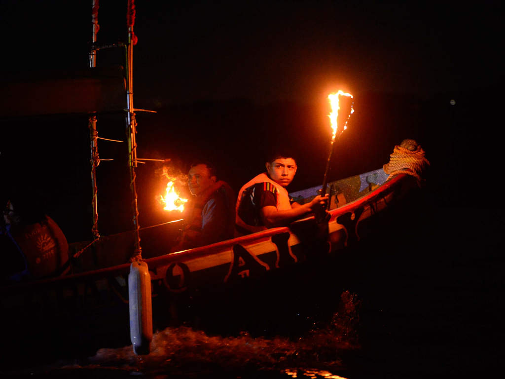 Dos personas en una lancha pequeña y oscura en el lago por la noche. La persona detrás sostiene una antorcha encendida que proyecta un brillo cálido. Llevan chalecos salvavidas. El agua está oscura.