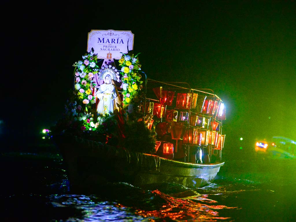 Una toma nocturna a media distancia de la lancha que transporta la estatua de la Virgen de los Dolores en el lago de Ilopango. La lancha está iluminada, mostrando las flores y el letrero "MARÍA, PRIMER SAGRARIO", rodeada por la oscuridad del lago y otras luces distantes.