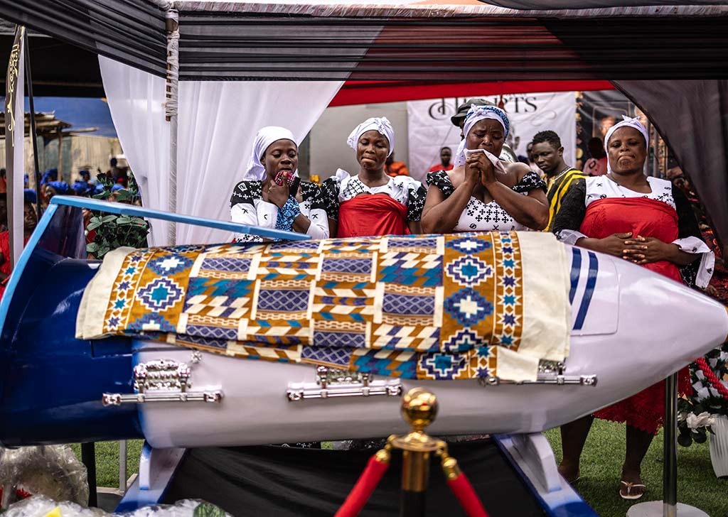 Cuatro mujeres vestidas con ropa tradicional blanca y pañuelos rojos en la cabeza lloran alrededor de un ataúd de madera blanco y azul con forma de avión. El ataúd está cubierto con una tela estampada de colores brillantes.