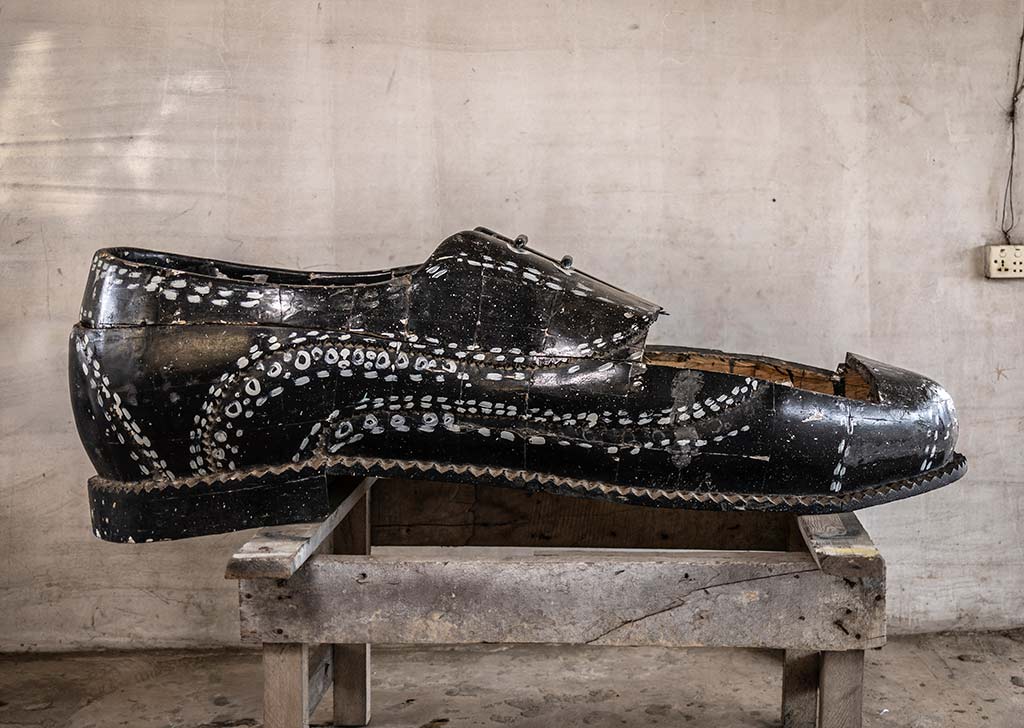 Un ataúd de madera con forma de zapato clásico de color negro con detalles intrincados de color blanco descansa sobre un soporte de madera. El ataúd está en un taller de carpintería.