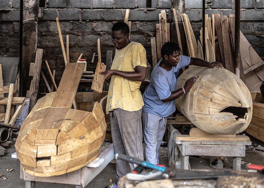 Dos jóvenes trabajan en un taller de carpintería, construyendo un ataúd de madera con forma de avión. Un niño lija la aleta de cola, que tiene la bandera de Ghana pintada. El otro niño está detrás, trabajando en el cuerpo del avión.