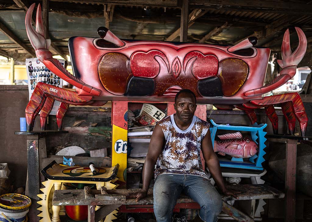 Un carpintero de Ghana se sienta frente a un gran ataúd de madera con forma de cangrejo de color rojo brillante en un taller de madera. El cangrejo tiene grandes pinzas y ojos detallados. Al fondo se ven otras tallas de madera y latas de pintura. El hombre lleva una camiseta sin mangas estampada.