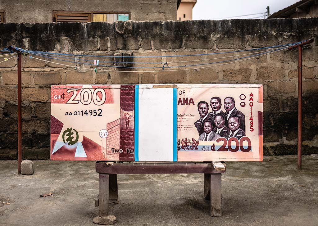 Un ataúd de madera grande con forma de billete de 200 cedis ghaneses descansa sobre un soporte de madera. El ataúd está pintado de naranja y blanco con el retrato de seis hombres en el frente.