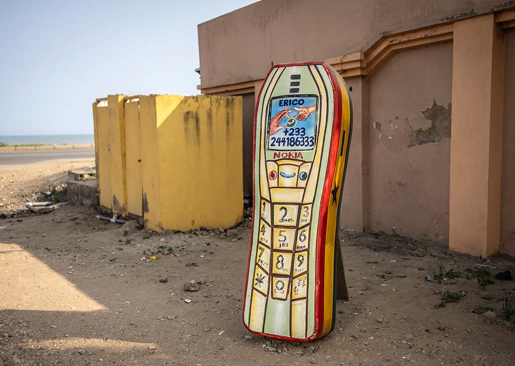 Un ataúd de madera con forma de teléfono móvil Nokia clásico de color amarillo y rojo descansa en la arena. El ataúd tiene botones de teléfono pintados y un número de teléfono en la pantalla. Al fondo, se ve el mar y el cielo azul.