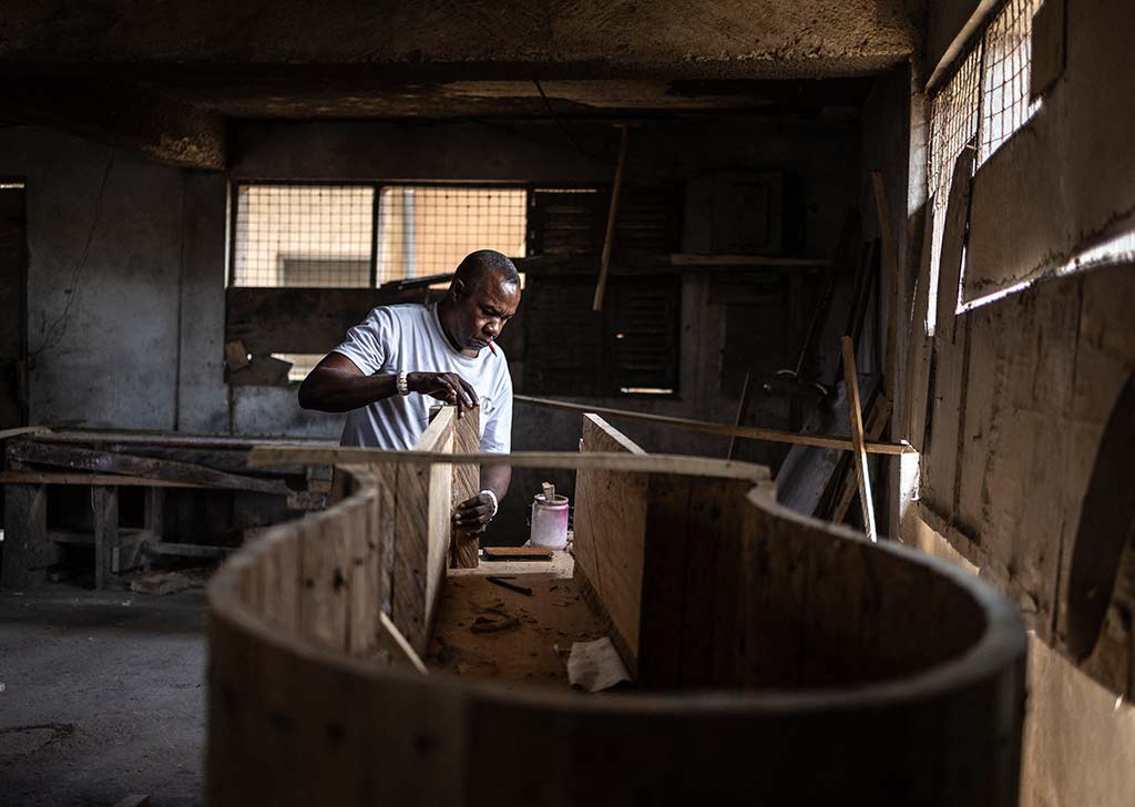 Un carpintero trabaja en un taller de carpintería, usando un cepillo de mano para dar forma a un ataúd de madera. El taller está oscuro y desordenado.