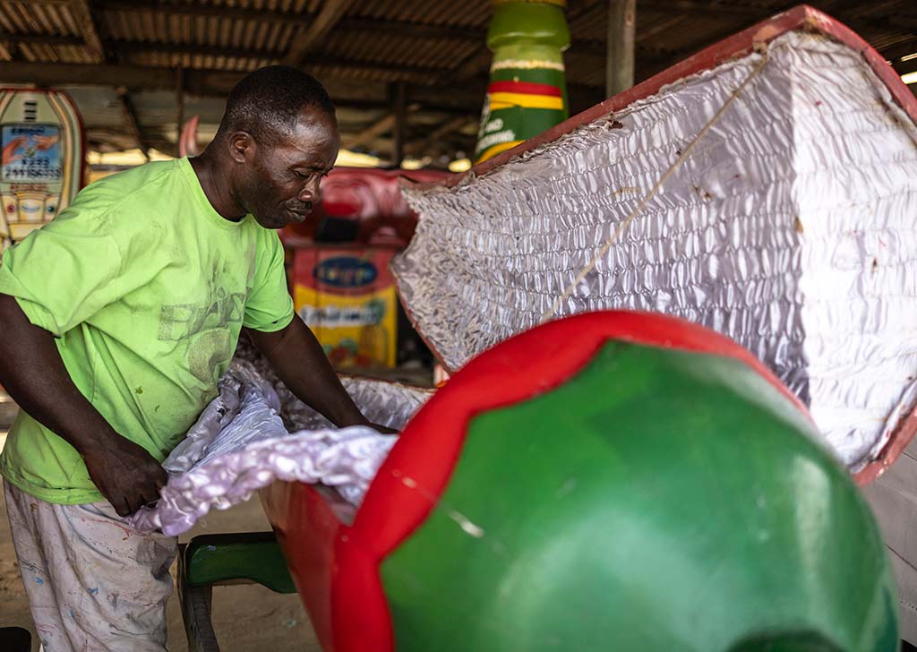 Un carpintero de Ghana en un taller de madera, inclinándose para pulir un gran ataúd de madera acolchado con forma de jeringuilla hipodérmica blanca y roja. El interior está forrado con una tela acolchada de color plateado. Se ven latas de pintura y herramientas al fondo.