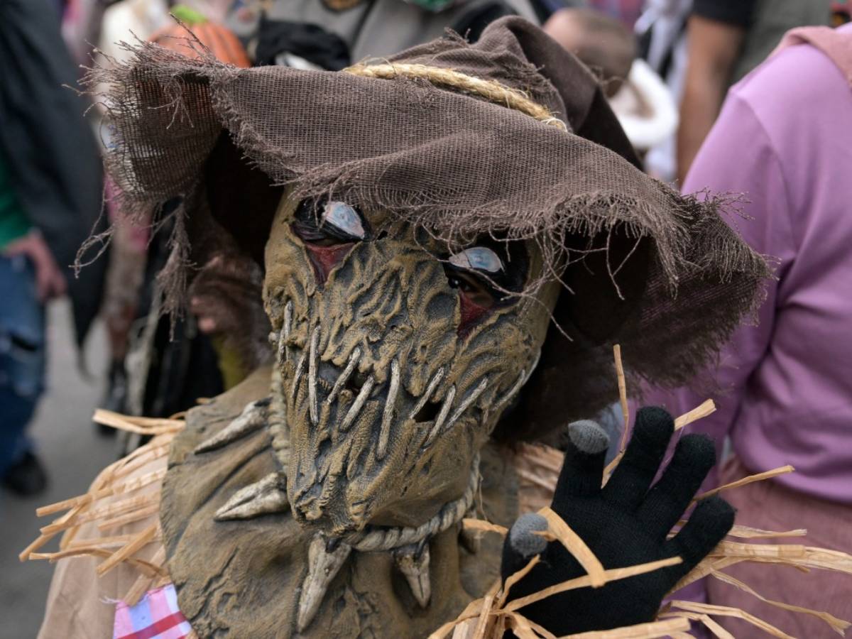 Persona disfrazada de espantapájaros macabro con sombrero de arpillera y guantes oscuros durante un desfile.