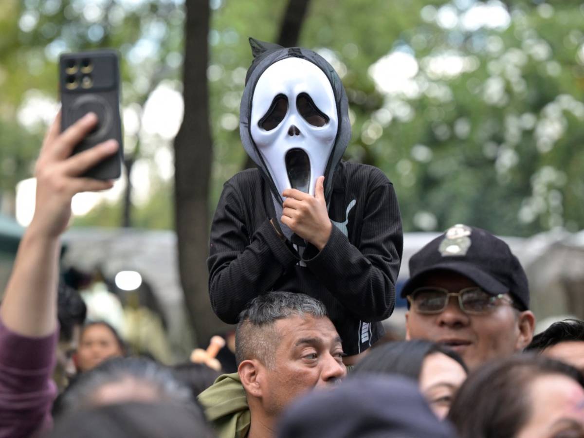 Niño disfrazado con la máscara de Ghostface sentado en hombros sobre la multitud en una calle arbolada.
