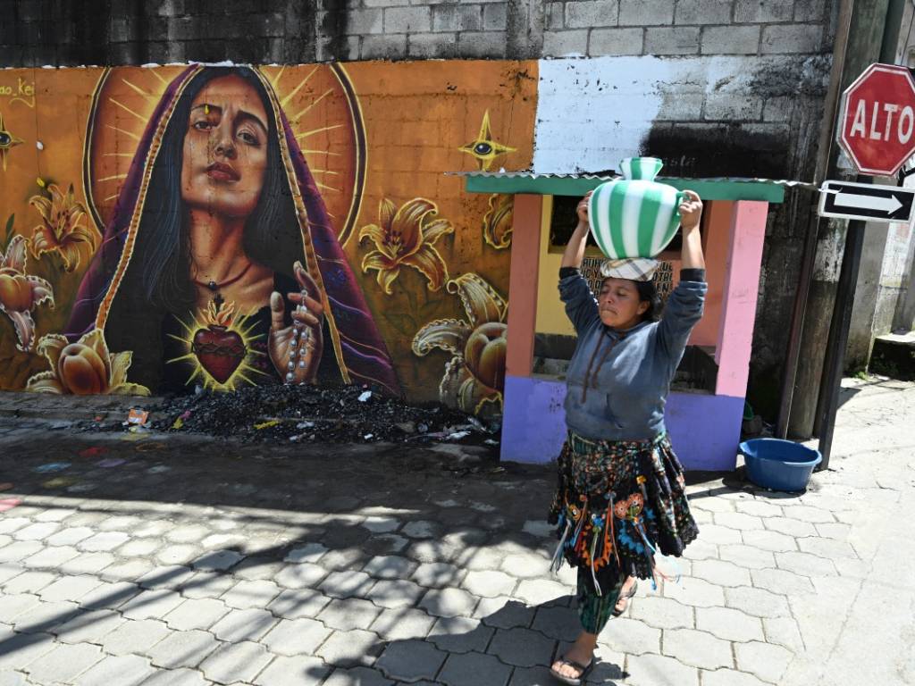Una mujer vestida con traje tradicional camina cargando un cántaro sobre la cabeza junto a un mural colorido de la Virgen del Sagrado Corazón de Jesús.