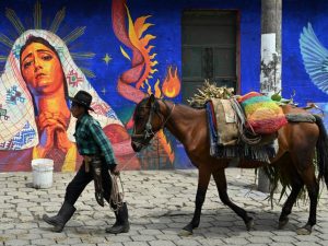 Un hombre indígena caminando junto a un caballo en una calle flanqueada por un mural religioso colorido en Guatemala.