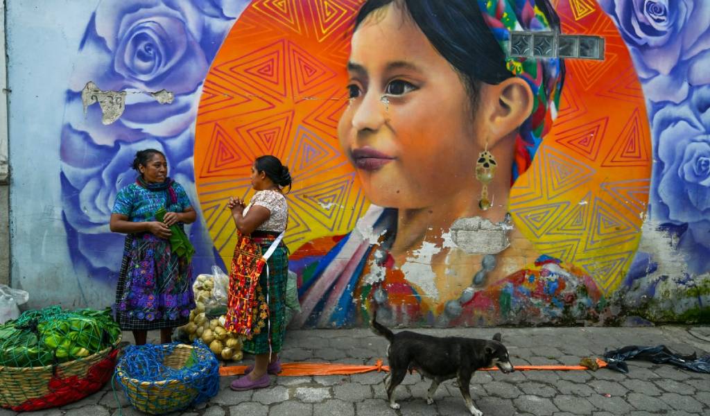 Dos mujeres indígenas vestidas con trajes tradicionales conversan de pie junto a un mural colorido que representa rostros indígenas.