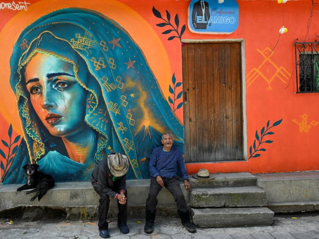 Dos personas con vestimenta tradicional descansan sentadas frente a un mural colorido de la Virgen de los Dolores en la calle.