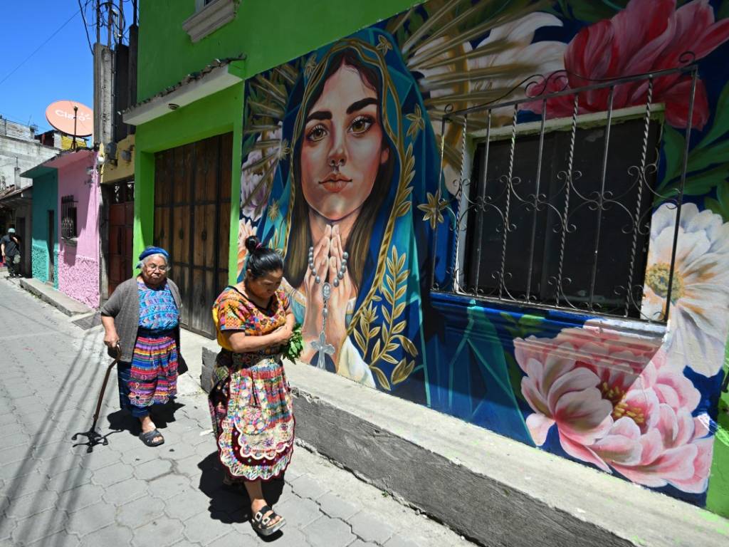 Una mujer indígena vestida con traje tradicional camina por una calle junto a un mural colorido de la Virgen María.