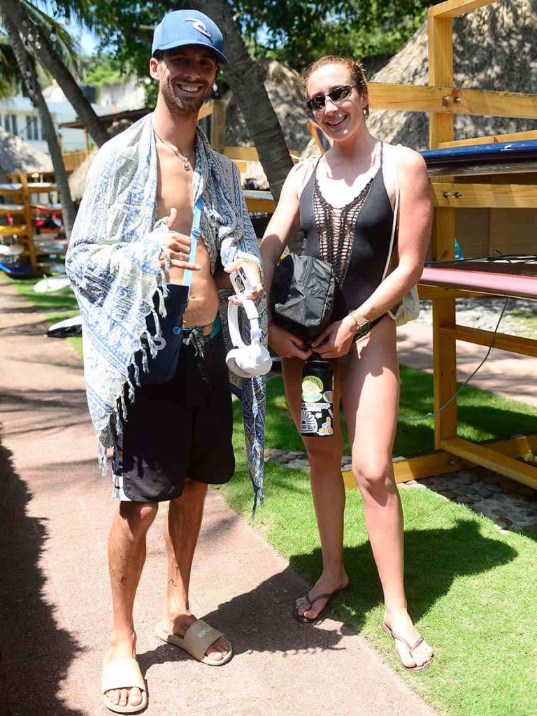 Pareja de surfistas luciendo toalla y audífonos posando junto al rack de tablas de surf.
