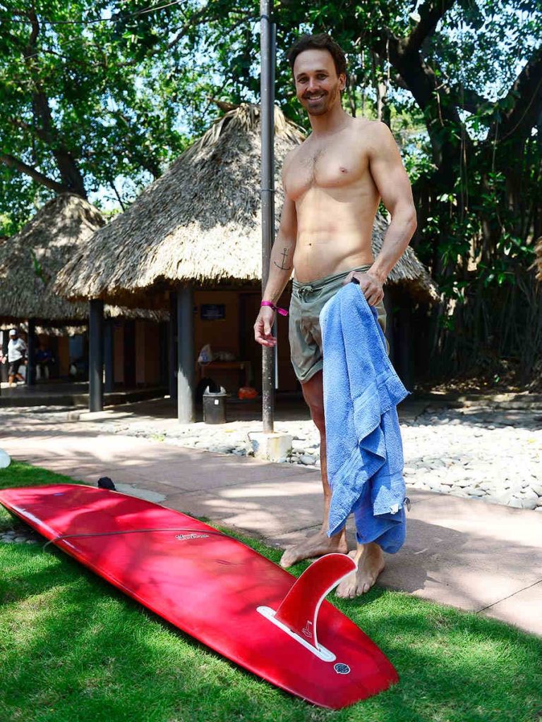 Surfista sosteniendo una toalla azul junto a su tabla larga roja con quilla sobre el césped.