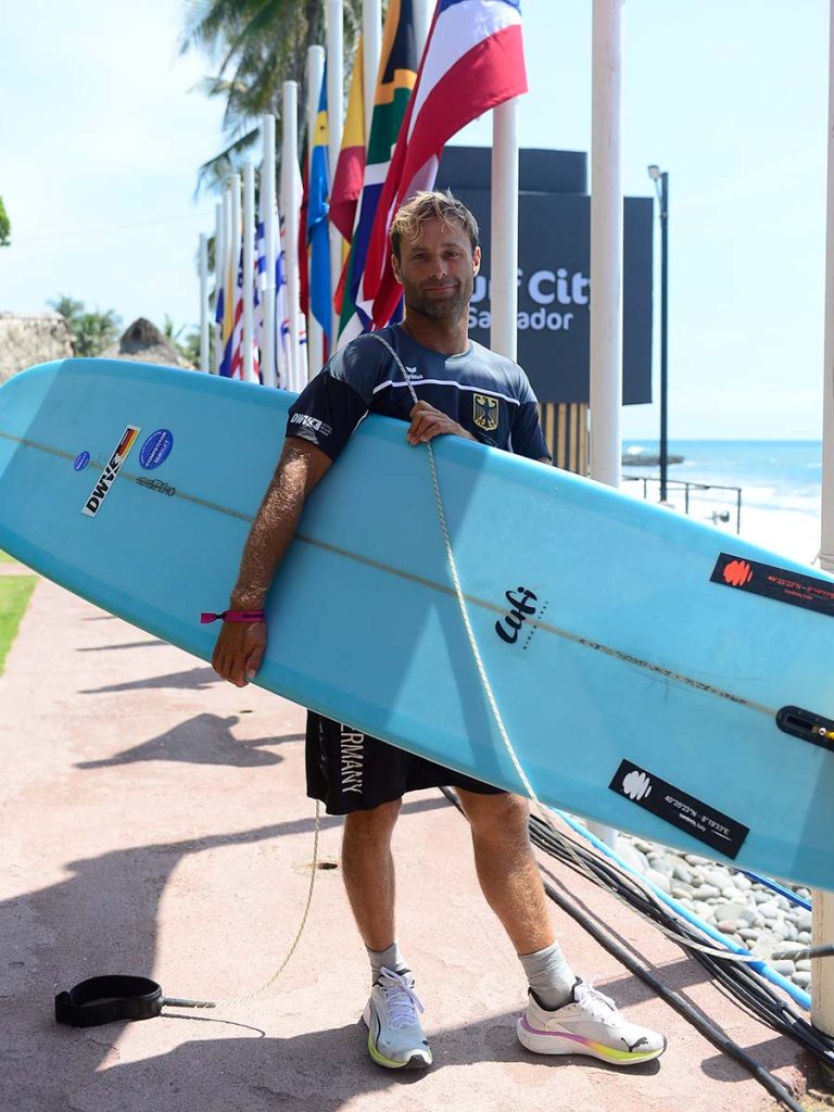 Competidor del equipo de Alemania sosteniendo su tabla de longboard celeste frente a las banderas oficiales del evento.