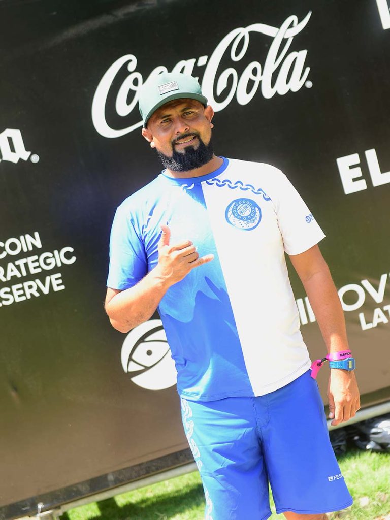 Entrenador de la selección salvadoreña de surf haciendo la señal del "shaka" frente al panel publicitario de Surf City.