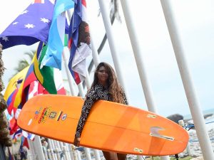 Surfista sosteniendo una tabla larga anaranjada mientras posa junto a la hilera de banderas de los países participantes.