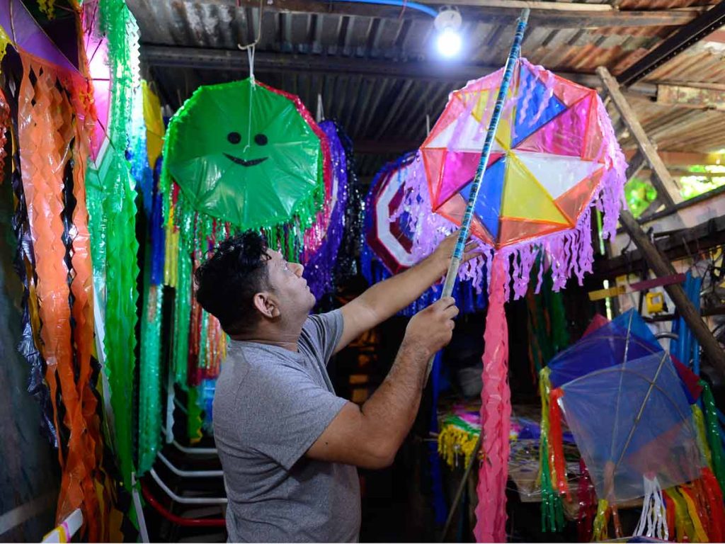 Hombre ajustando con una vara una piscucha multicolor colgada del techo del taller entre decenas de cometas.