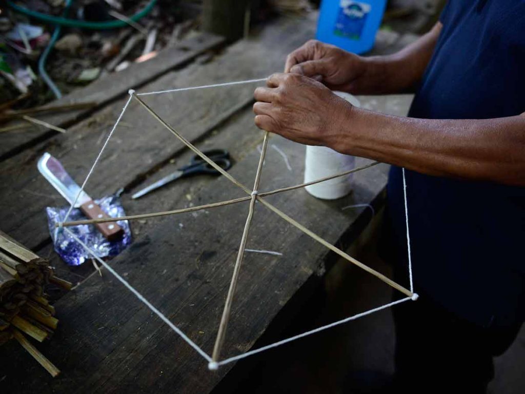Manos de Don Ramiro uniendo y amarrando con hilo blanco el centro y los extremos de una estructura hexagonal de bambú