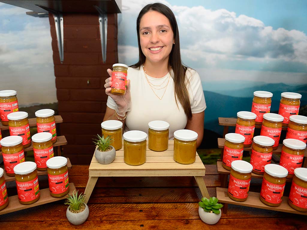 Mujer sonriente posando junto a su colección de frascos de conservas y jaleas artesanales de jocote.