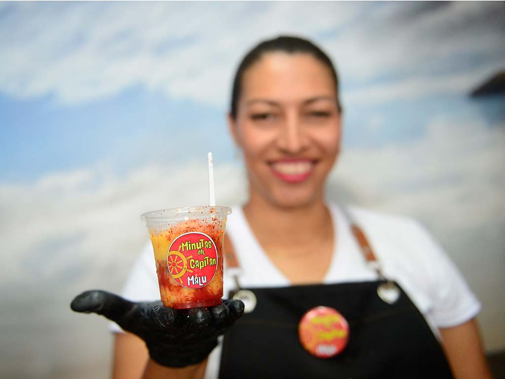 Vaso transparente con minuta helada cubierta de chilitos y jarabe artesanal de fruta sostenido por una emprendedora.