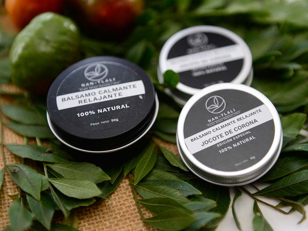 Envases metálicos de bálsamo relajante 100% natural de jocote de corona colocados sobre hojas verdes.