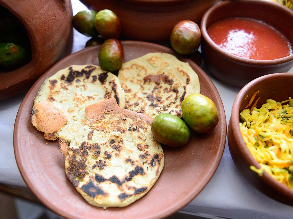 Plato de barro con pupusas recién hechas, jocotes frescos verdes y cuencos con salsa y curtido.