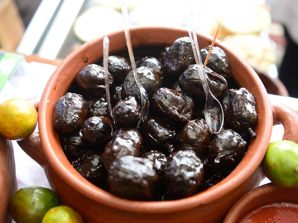 Cazuela de barro llena de jocotes en miel acaramelados con utensilios para degustación.