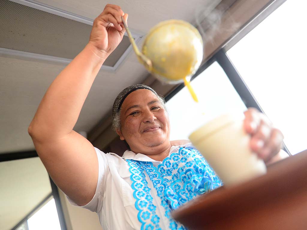 Mujer con vestimenta tradicional sirviendo atol de jocote humeante con un cucharón en vaso térmico.