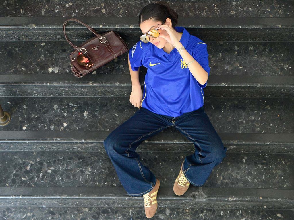 Cantante salvadoreña Gabriela Triste sentada en unas escaleras luciendo lentes amarillos y camisa azul del Chelsea.