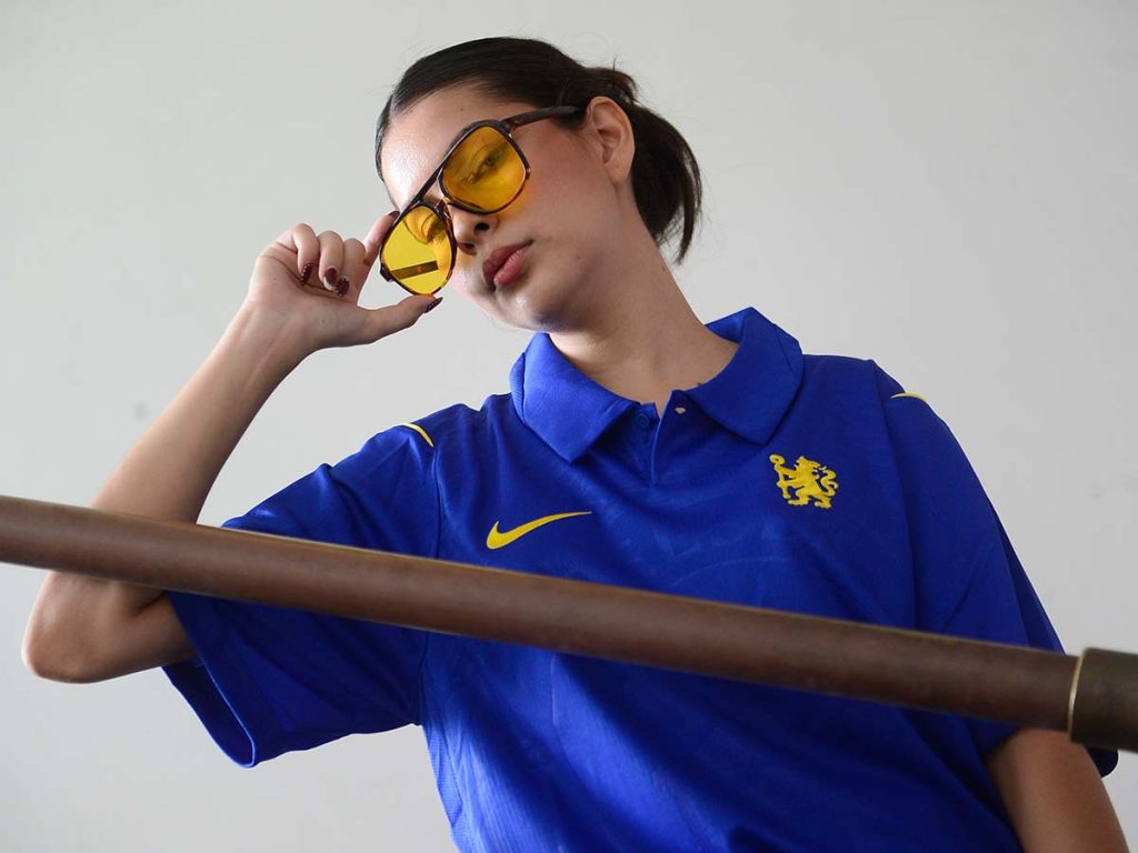 Gabriela Triste posando apoyada en un pasamanos de metal con gafas oscuras y camisa deportiva.