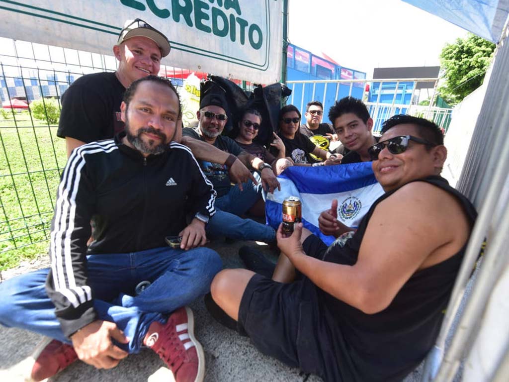 Grupo de fanáticos sentados en el suelo descansando a la sombra junto a una valla metálica.