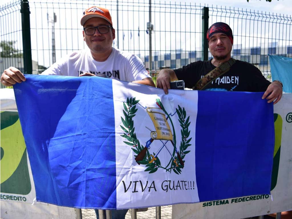 Dos jóvenes extendiendo la bandera azul y blanco de Guatemala en la acera del recinto.