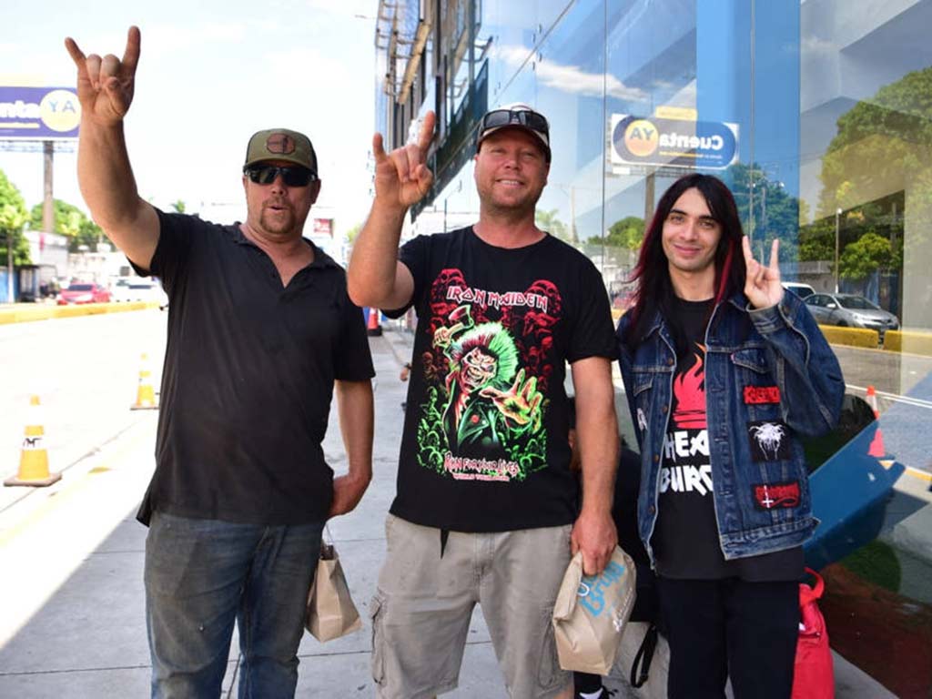 Tres seguidores de la banda de metal posando en la acera; dos hombres alzando la mano con el gesto del rock y un joven haciendo la señal de la paz.
