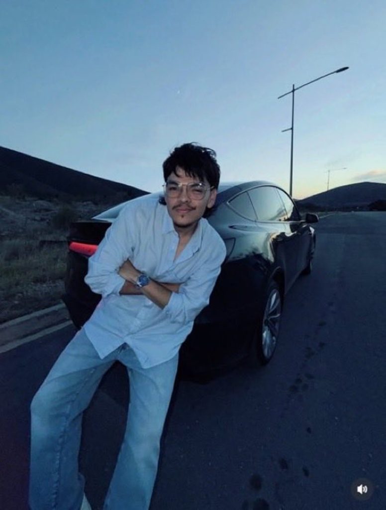 Streamer El Komanche apoyado sobre la parte trasera de su auto Tesla color negro en una carretera al atardecer.