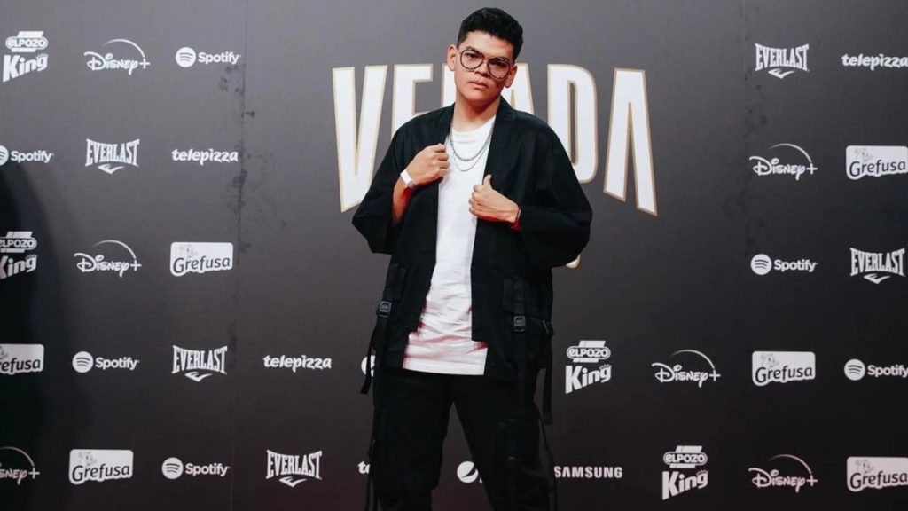 El Komanche posando en alfombra roja de evento masivo de streaming con kimono negro y fondo publicitario.