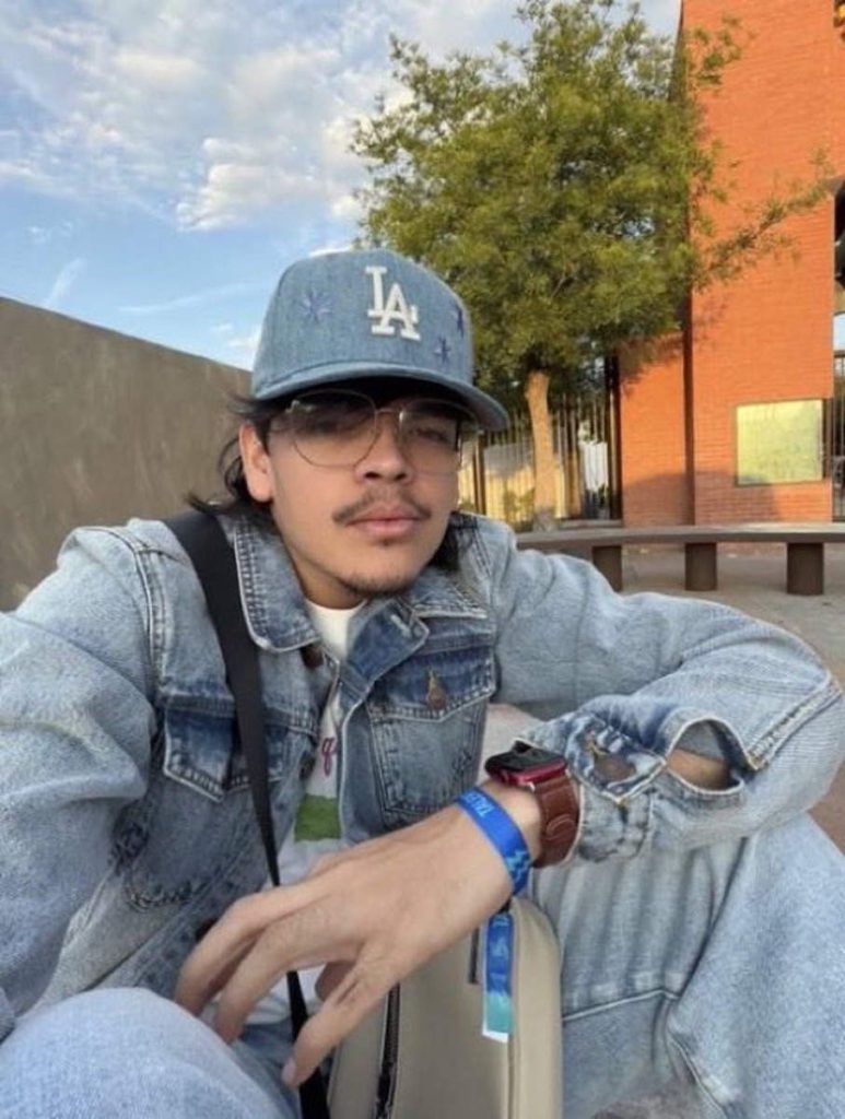 El Komanche sentado al aire libre luciendo gorra azul de los Dodgers, chaqueta de jeans y anteojos.