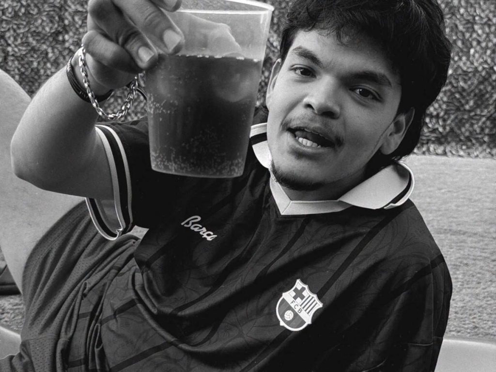 Fotografía en blanco y negro de El Komanche sonriendo y sosteniendo un vaso para brindar con camiseta deportiva.