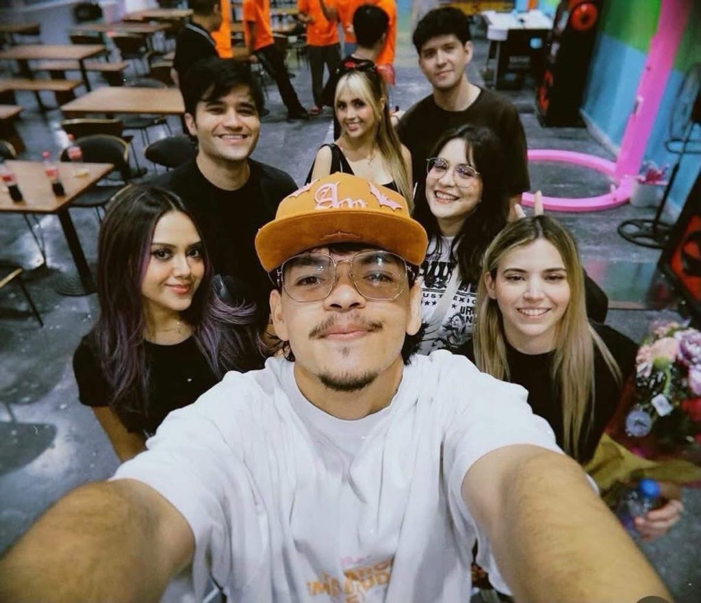 Selfie tomada por El Komanche con gorra anaranjada junto a un grupo de amigas y streamers sonriendo.