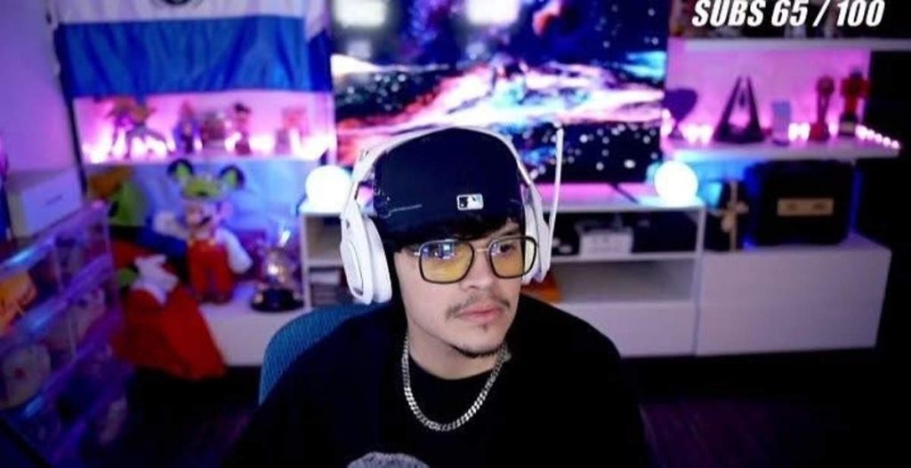 El Komanche con audífonos blancos, gorra negra y bandera de El Salvador al fondo durante un stream.