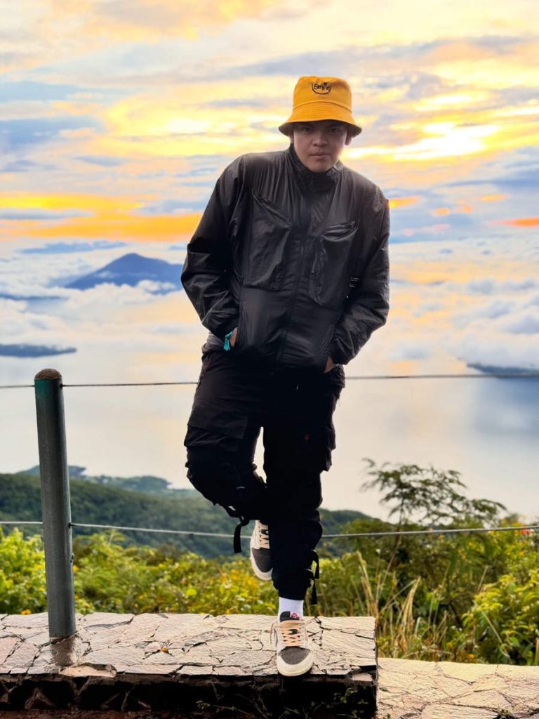 El Komanche con sombrero amarillo y chaqueta negra posando en un mirador con vistas a un lago y montañas.
