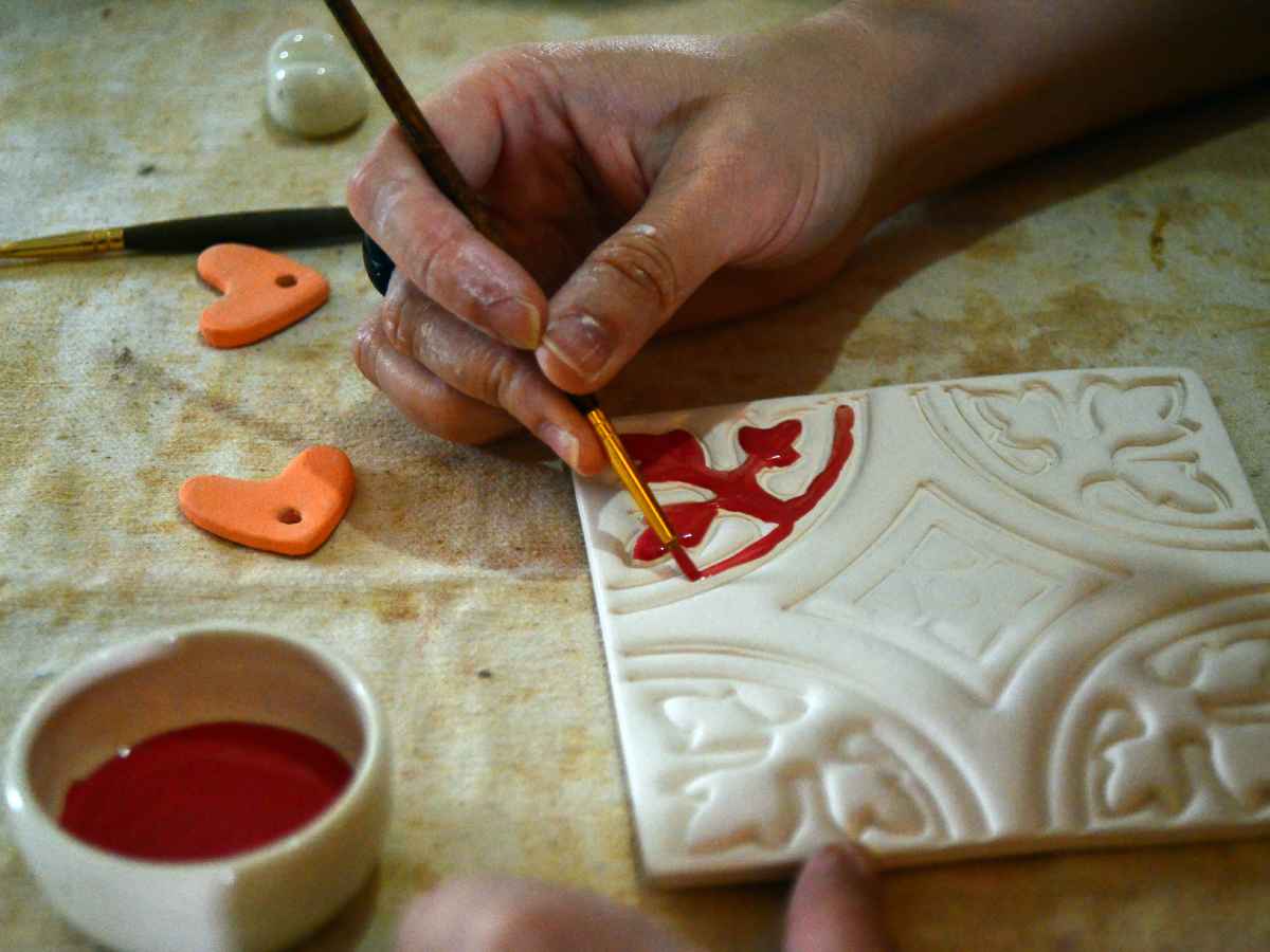 Fotografía en primer plano de manos artesanas pintando con pincel fino los relieves de un azulejo de cerámica blanca.