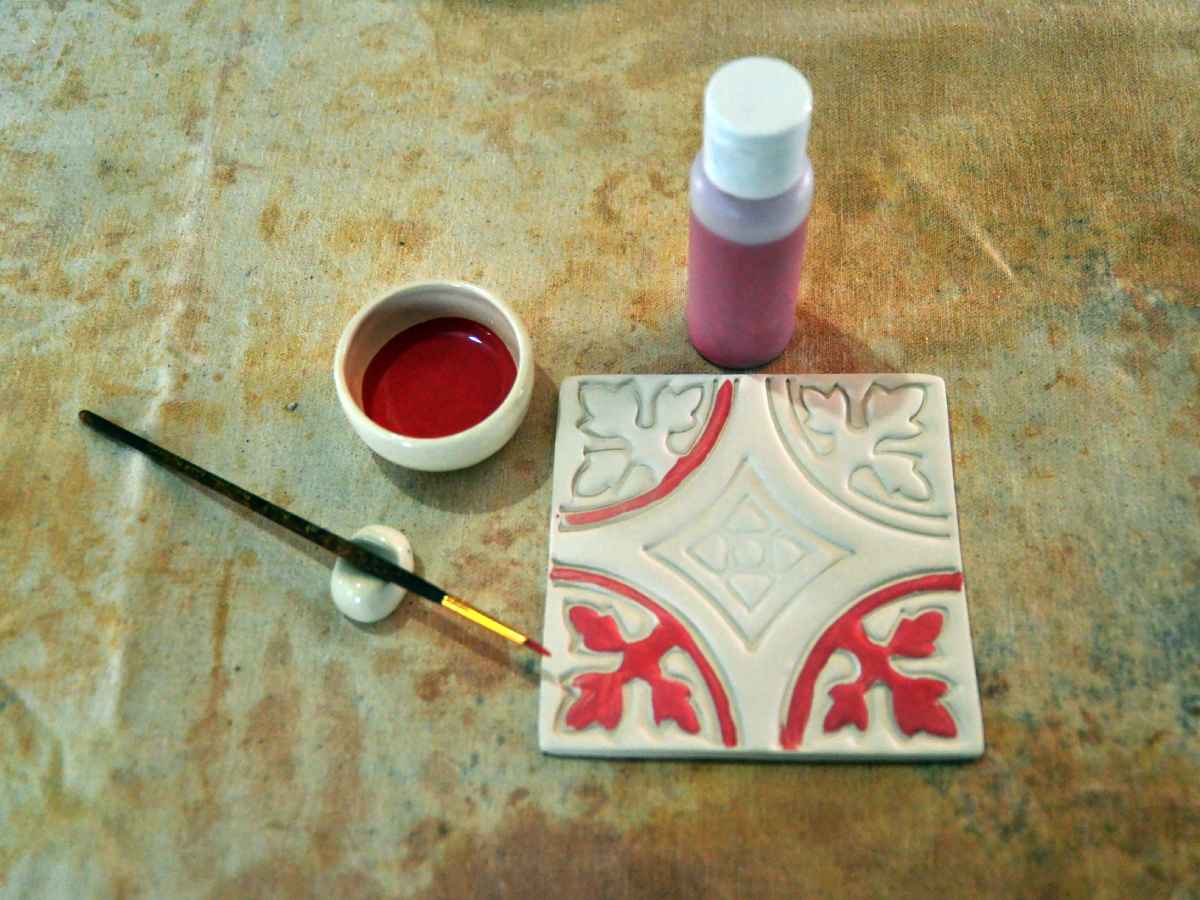 Fotografía cenital de un azulejo de cerámica con detalles en pigmento rojo, acompañado de un pincel fino, recipiente de pintura y frasco.