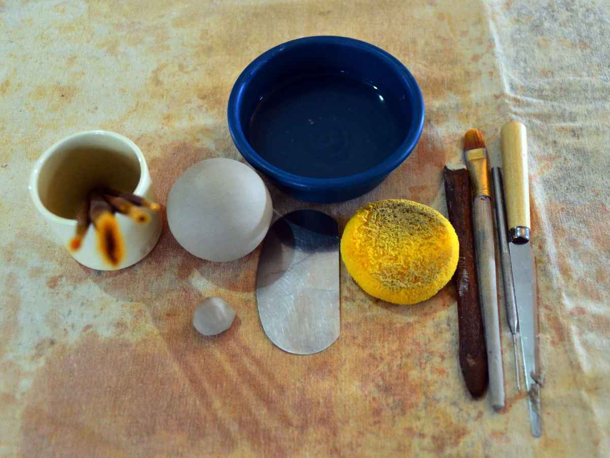 Fotografía cenital que muestra herramientas de alfarería como pinceles, estiletes, esponja amarilla, agua y masa de arcilla fresca.