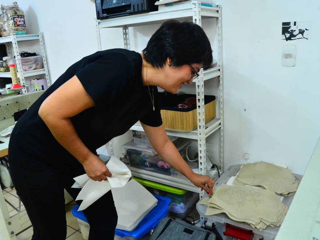 Nancy Herrera se inclina en su taller para tomar piezas de tela dibujadas agrupadas sobre una superficie cerca de la máquina.