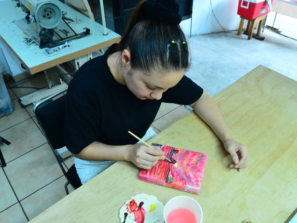 Gisela Herrera mezclando pintura acrílica sobre una paleta de arte frente a un lienzo de paisaje.