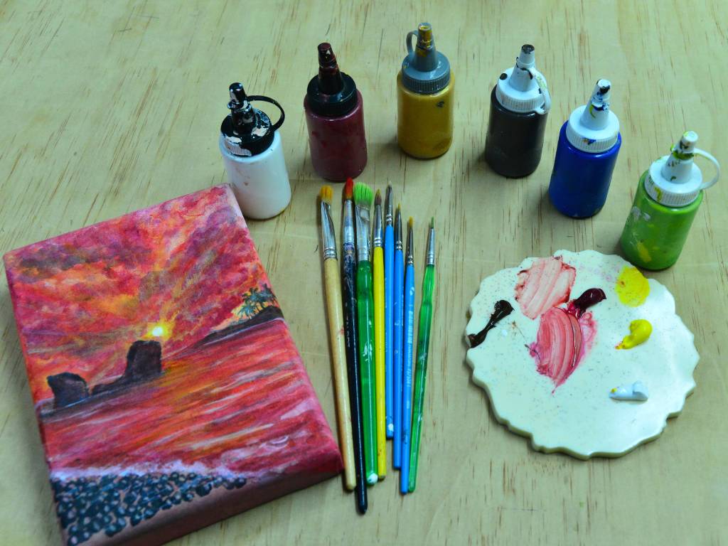 Diversos materiales de pintura, como pinceles, paleta con colores, tubos de acrílico y lienzos sobre la mesa de trabajo.